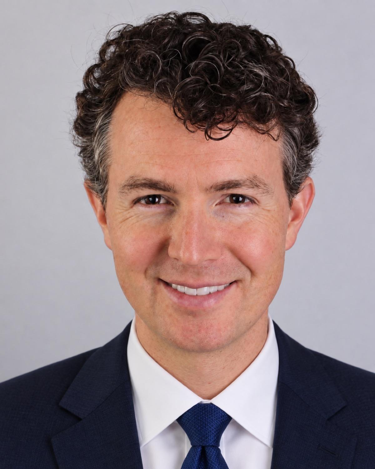 Dan Wood, head and shoulders, in a dark grey suit, white shirt and blue patterned tie, against a plain light grey background